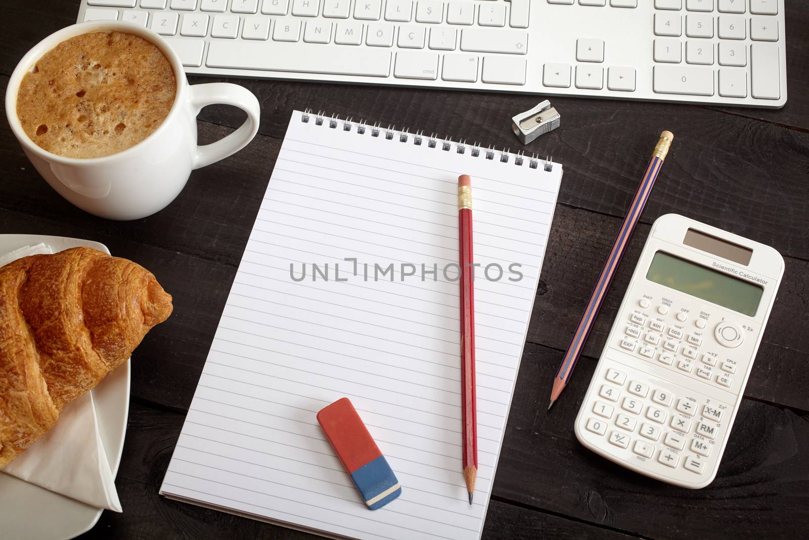 Top view office workplace - coffee, keyboard, pencil, croissant, calculator and notebook on black table