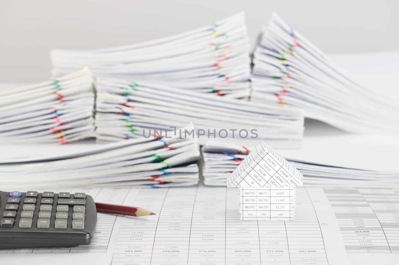 Brown pencil with calculator and house on finance account have blur pile overload paperwork of report and receipt with colorful paperclip place on white background.