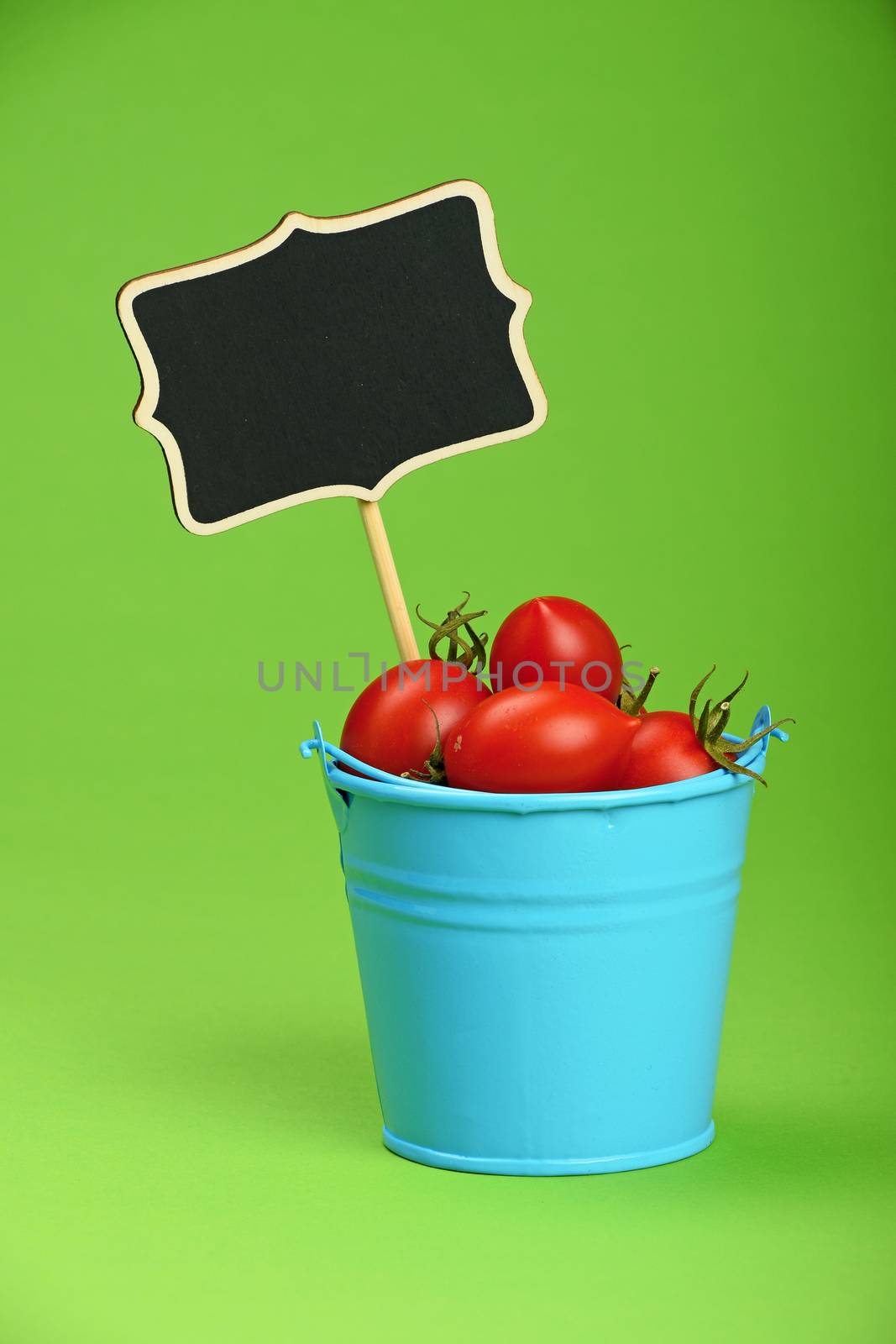Small disproportional blue bucket of red cherry tomatoes and black chalkboard sign over green background, close up
