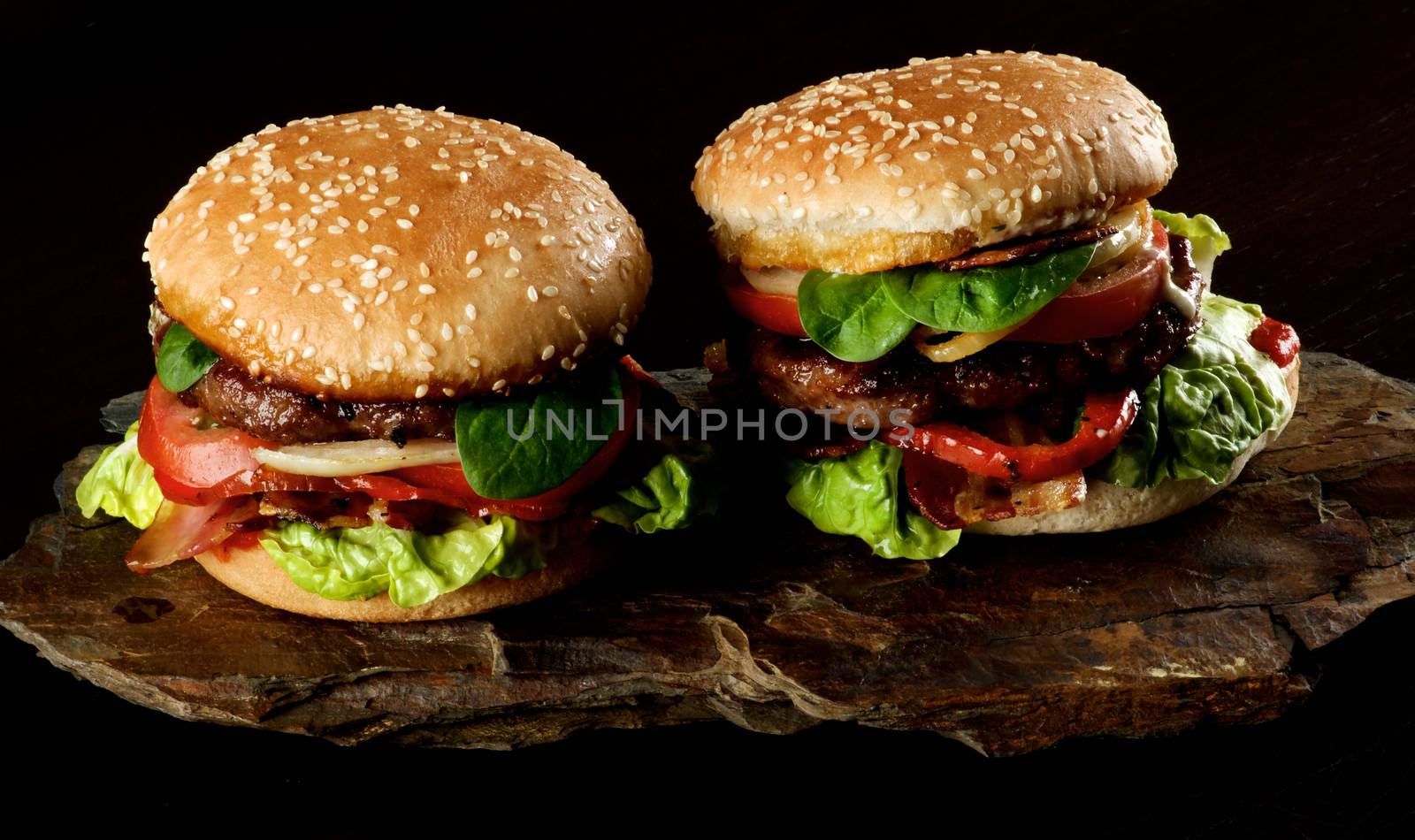 Two Tasty Hamburgers with Beef, Bacon, Lettuce, Tomatoes, Basil, Roasted Onion and Juicy Sauce on Sesame Buns on Stone Board closeup on Dark background