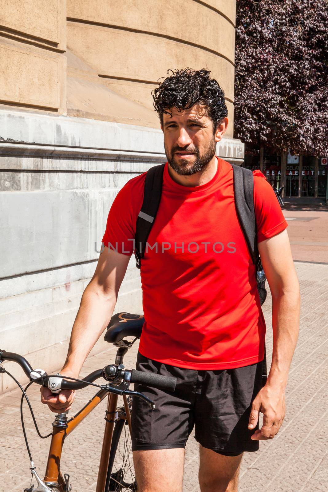 Urban athlete walking with a urban bike he is dressed with a red t-shirt a bag and black short sport trousers
