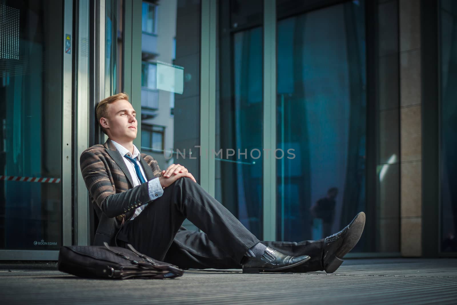 Young handsome successful stylish businessman sitting near modern office building. Concept: failure.