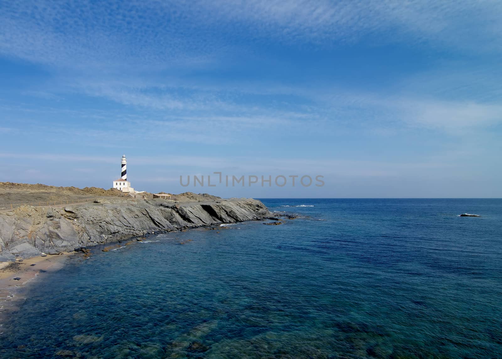 Lighthouse Cap de Favatrix with Rocky Shake Shore on Sea Waves Edge and Blue Sky background Still Life. Northern Coast of Menorca, Balearic Islands