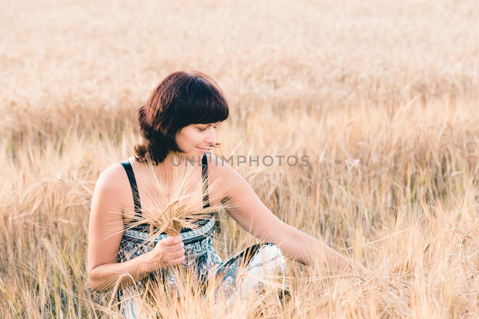 Middle aged beauty woman in a summer dress with no makeup relaxing in countryside tearing into bouquet of golden barley with her hand, summer concept