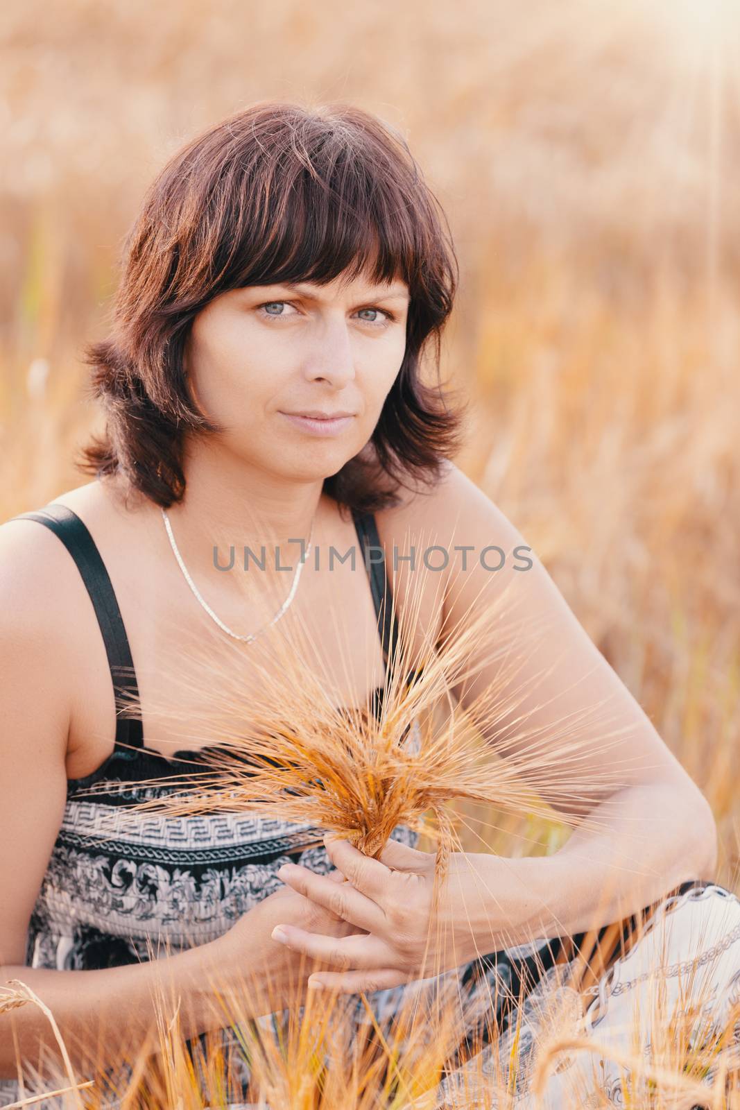 Middle aged beauty woman in a summer dress with no makeup relaxing in countryside tearing into bouquet of golden barley with her hand, summer concept