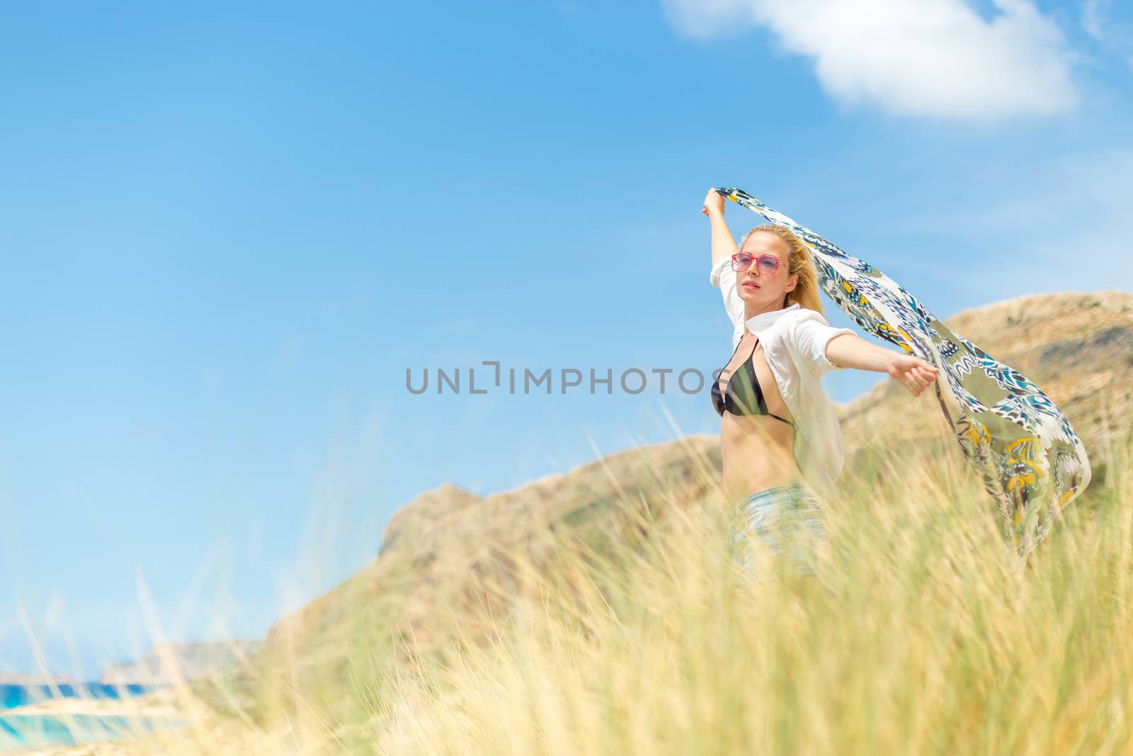 Relaxed woman, arms rised, holding colorful scarf, enjoying sun, freedom and life at beautiful beach. Young lady feeling free, relaxed and happy. Concept of vacations, freedom, joy and well being.