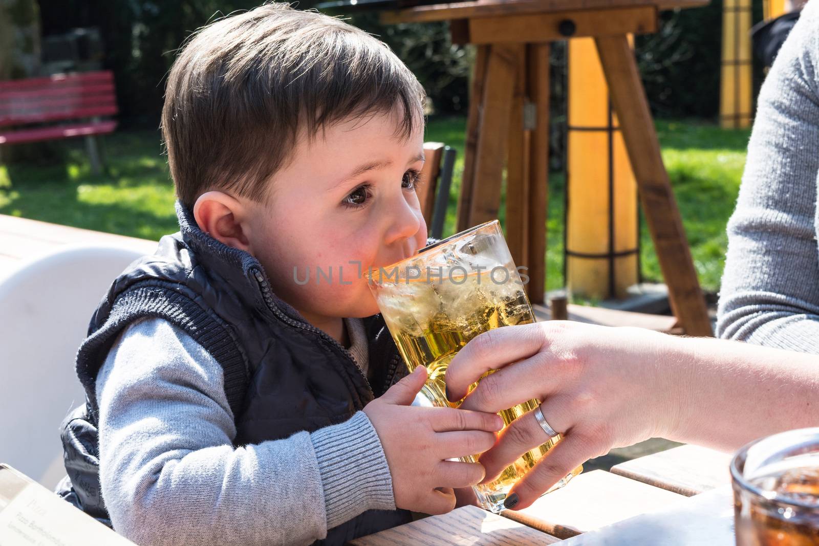 Child drinks juice from a glass in a garden restaurant.