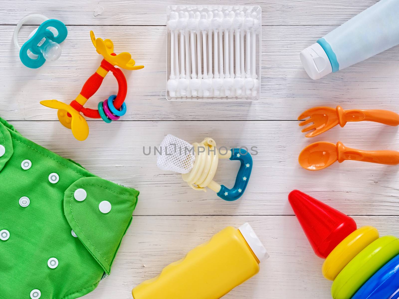 Collection of items for babies: cloth diaper, baby powder, nibbler, cream, teether, soother, cotton swabs, baby spoon and fork, pyramid toy on white wooden background. Top view or flat lay