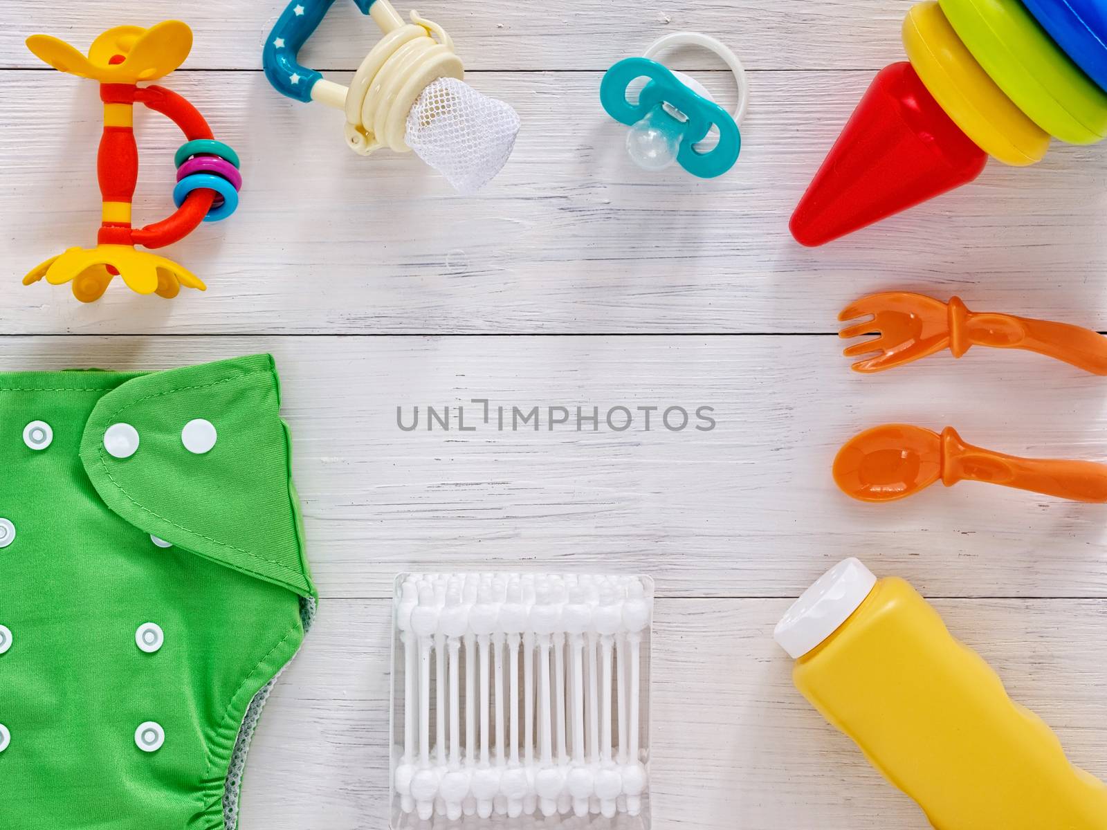 Babies goods: cloth diaper, baby powder, nibbler, teether, soother, cotton swabs, baby spoon and fork, pyramid toy on white wooden background with copy space. Top view or flat lay