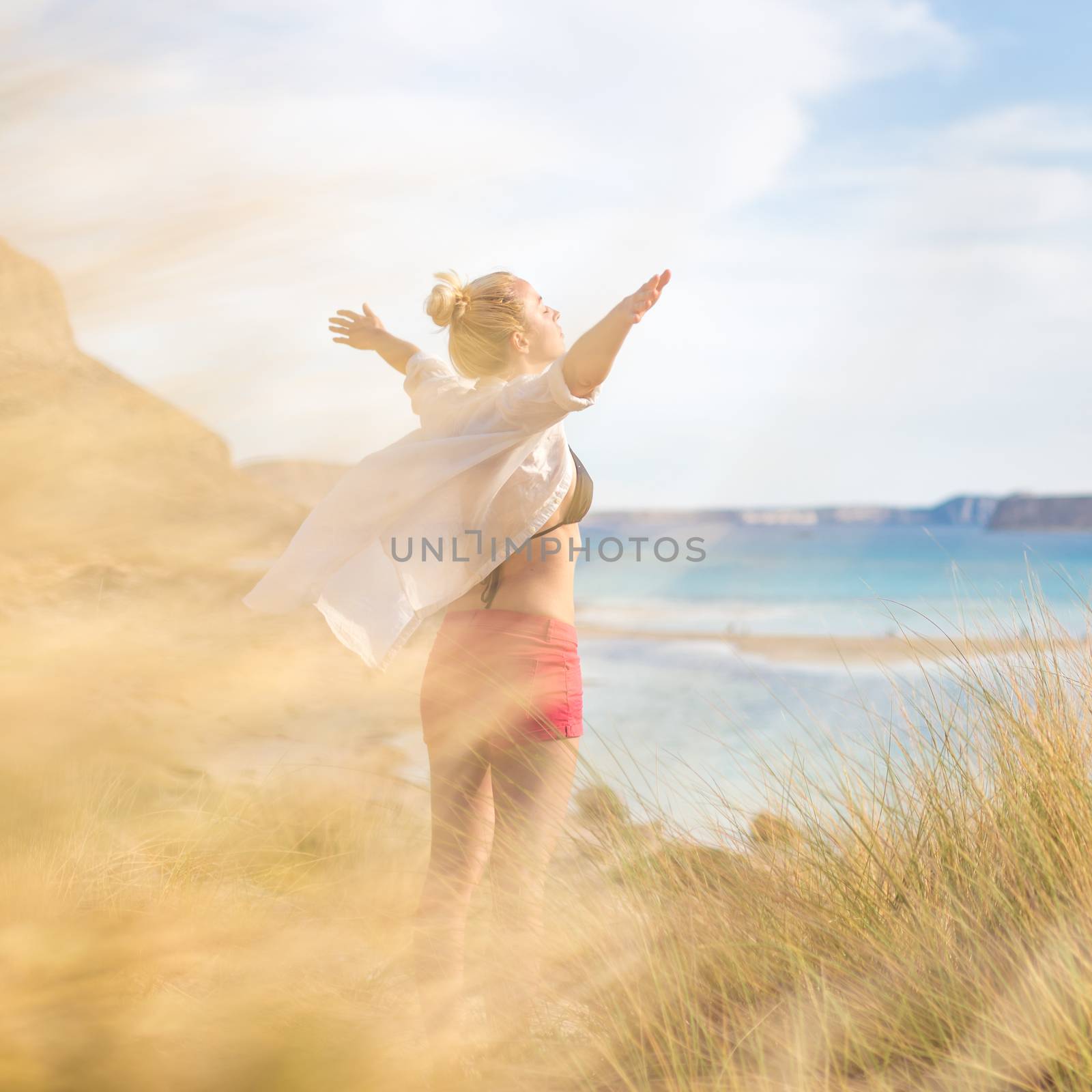 Relaxed woman in white shirt, arms rised, enjoying sun, freedom and life an beautiful beach. Young lady feeling free, relaxed and happy. Concept of vacations, freedom, happiness, joy and well being.