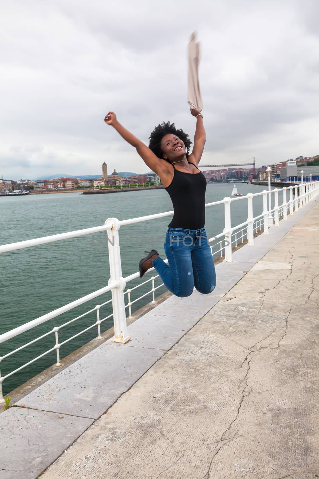 Beautiful African woman jumping near a boardwalk in a coastal city in a cloudy day