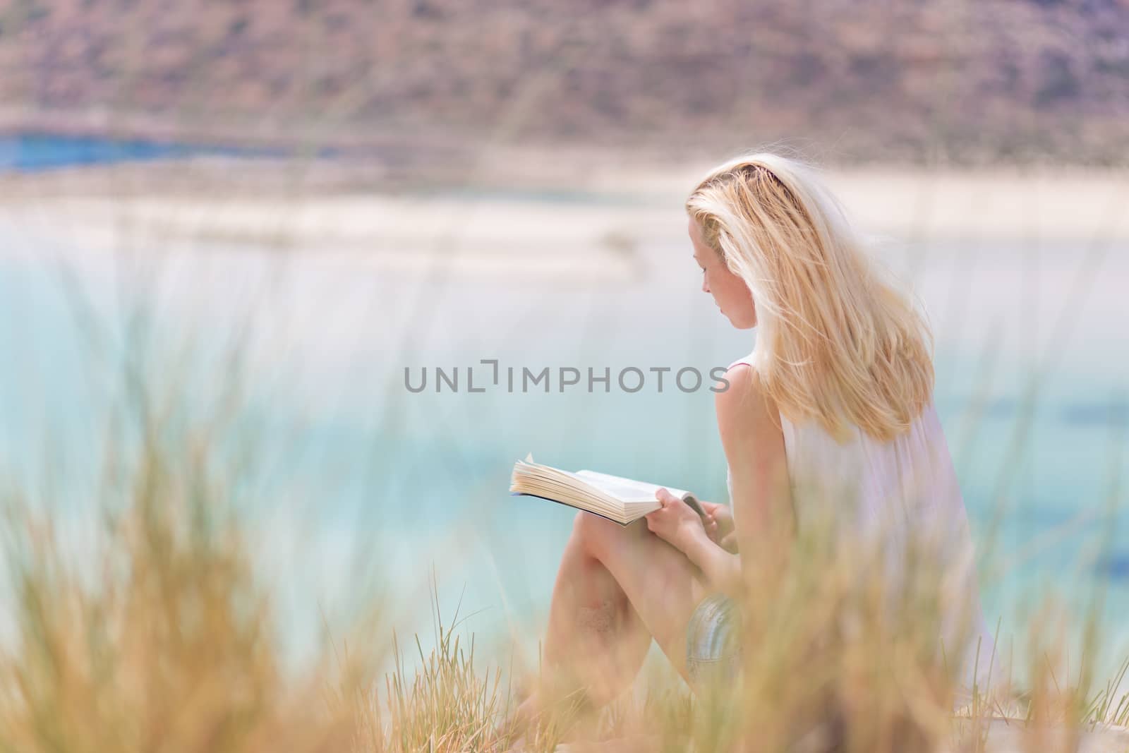 Relaxed woman enjoying sun, freedom and good book an beautiful sandy beach of Balos in Greece. Young lady reading, feeling free and relaxed. Vacations, freedom, happiness, enjoyment and well being.