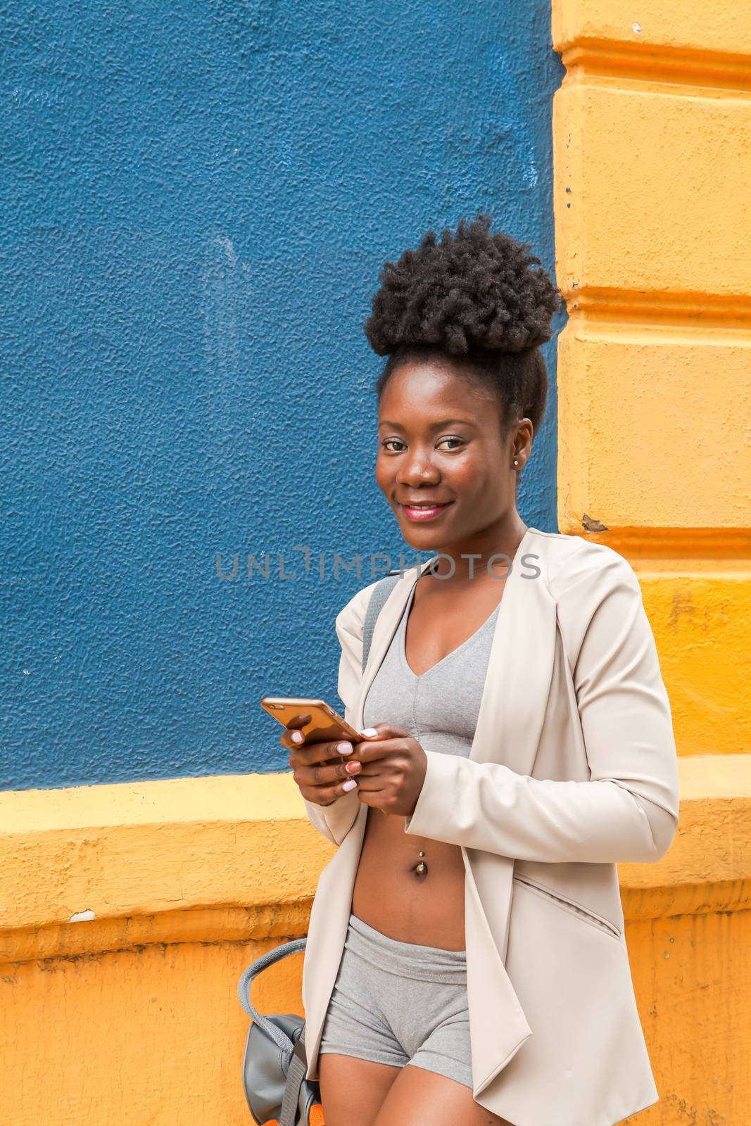 Medium shot of an african young woman in sportswear looking at his mobile phone with a colorful wall blue and yellow