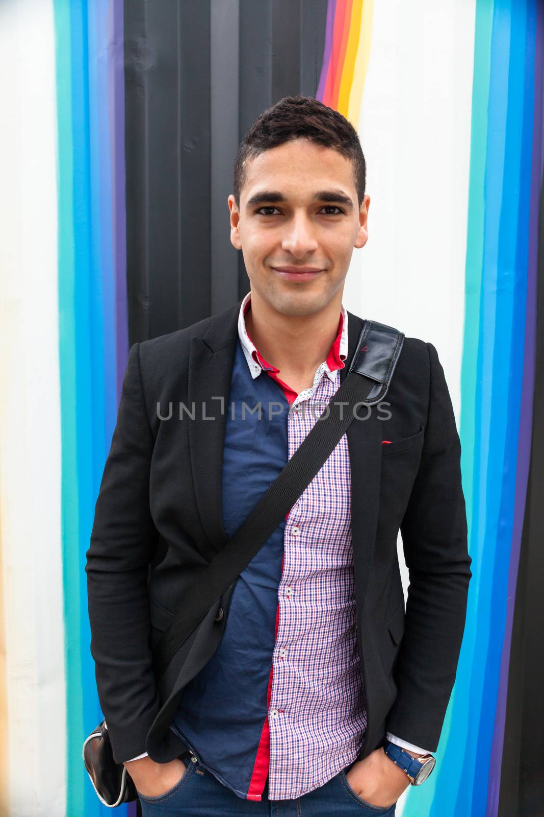 Modern young man with Arab features with shoulder bag and jacket without a tie on multicolored background and the hands in his pocket horizontal photography