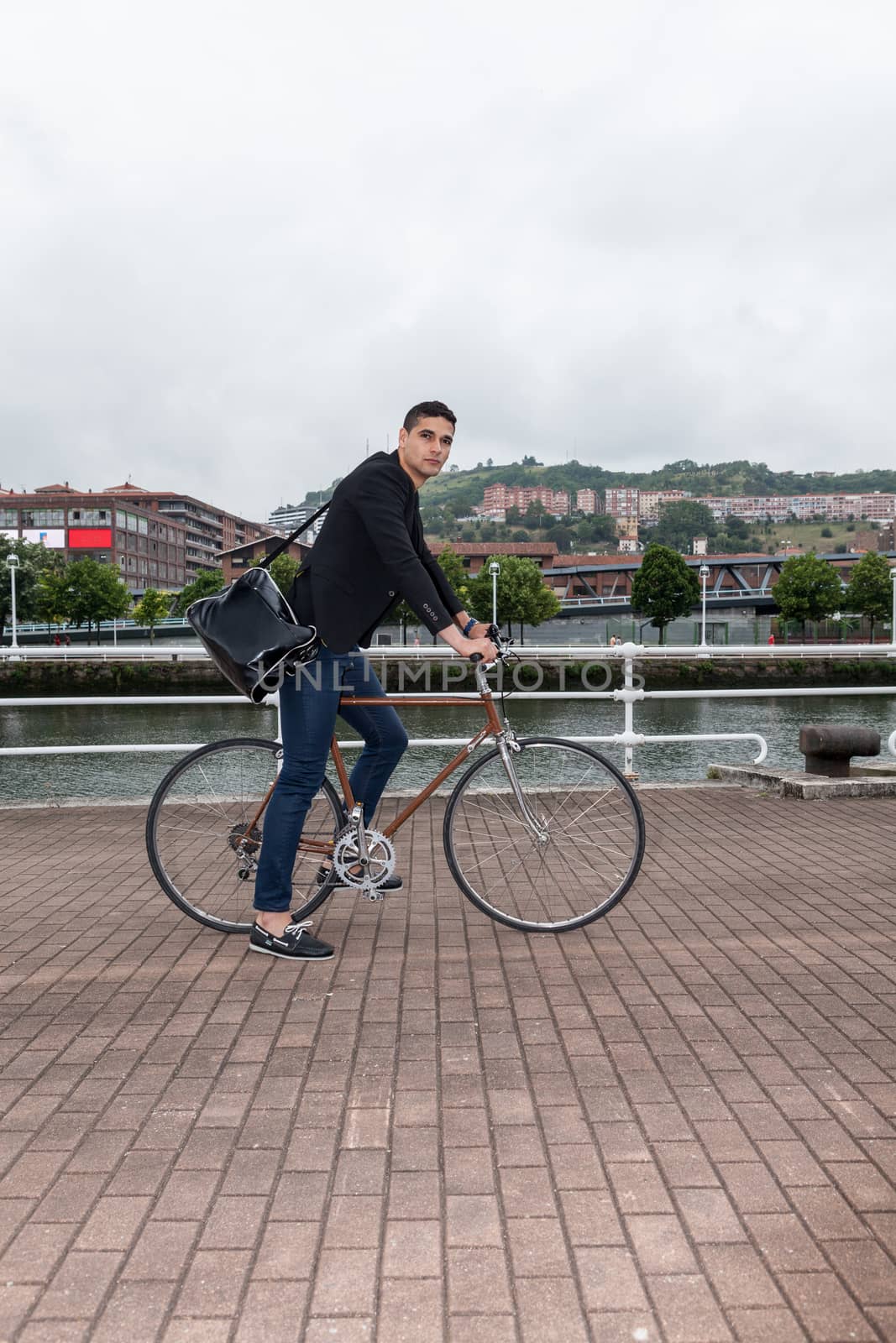 Close up or a young man riding on a bike on a cloudy day near the river with a shoulder bag and jeans on a cloudy day