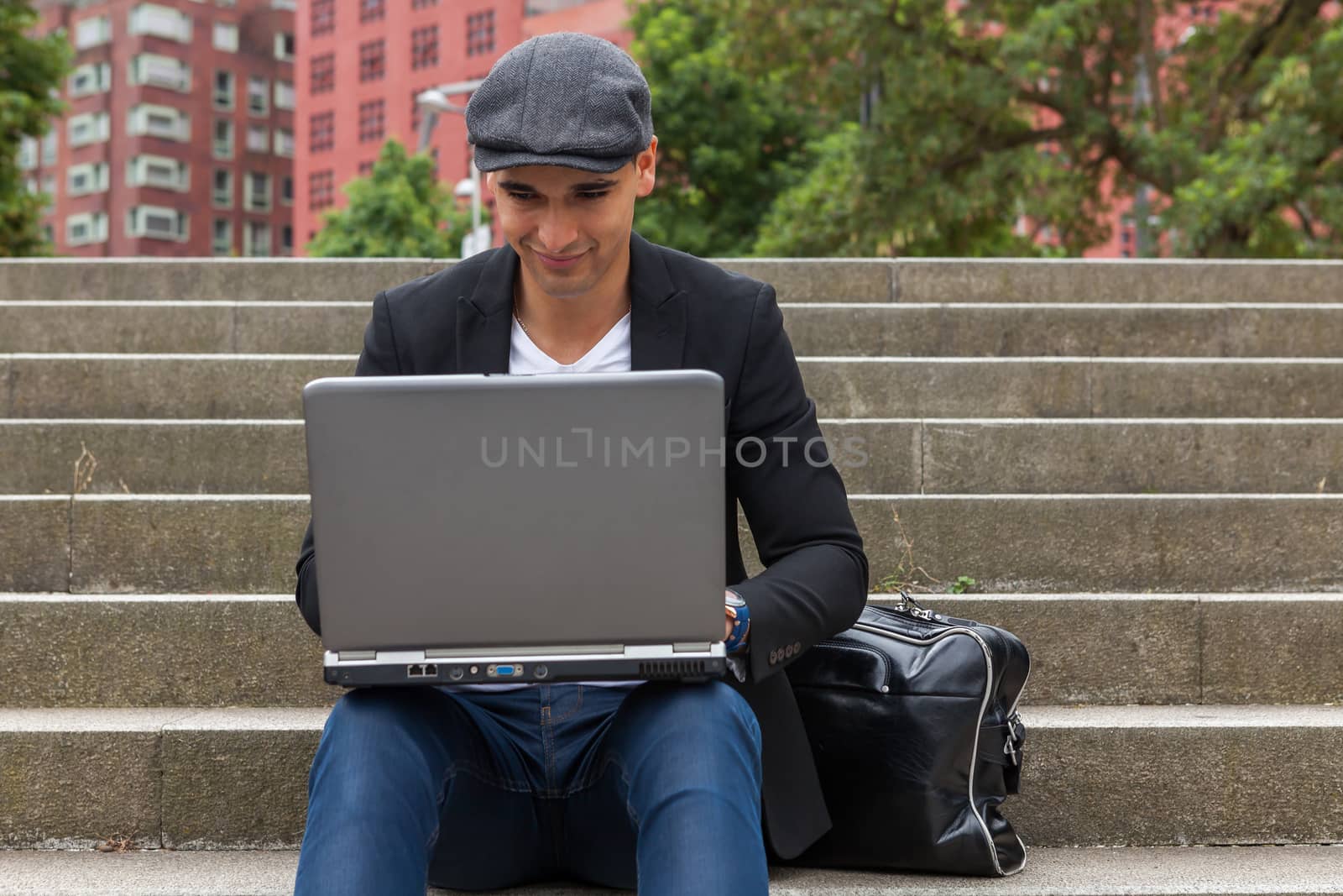 Young man with Irish beret sitting on a wall next to an urban stairs and a shoulder bag near
