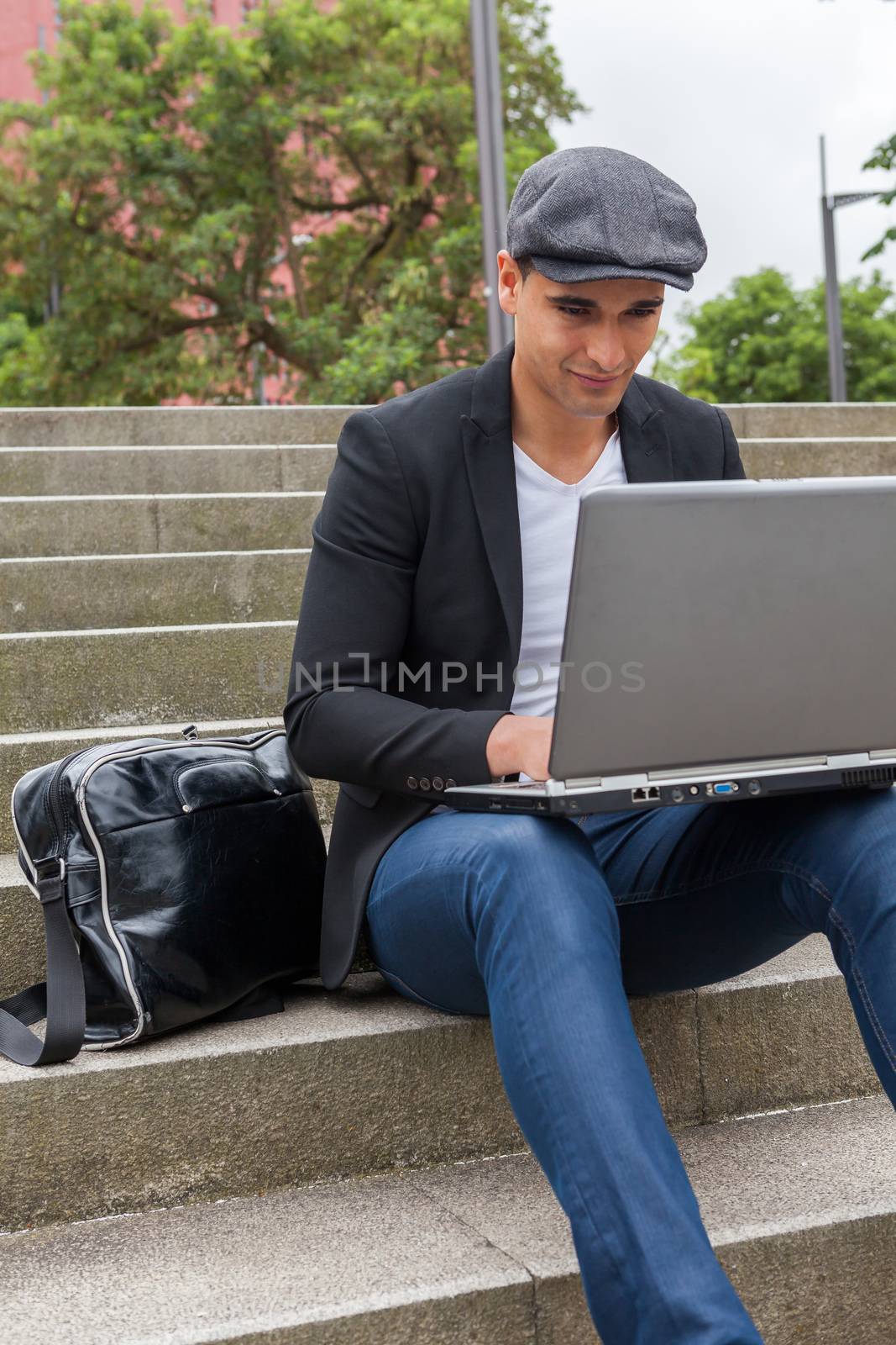 Young man with Irish beret sitting on a wall next to an urban stairs and a shoulder bag near
