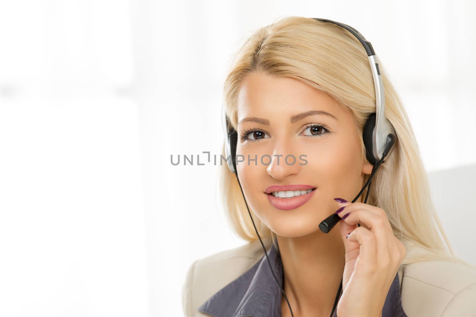 Close-up of a portrait young beautiful blonde girl, elegantly dressed, sitting at an office with a headset on her head. With a smile looking at the camera.
