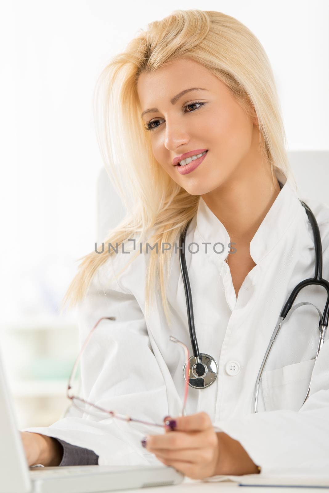 Attractive blonde female doctor in her consulting room, sitting at the table in front of laptop.