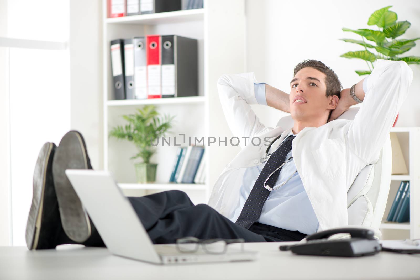 Young male successful doctor in the doctor's room sitting on chair, hands behind  head with her feet up on a desk. He is relaxing and daydreaming.