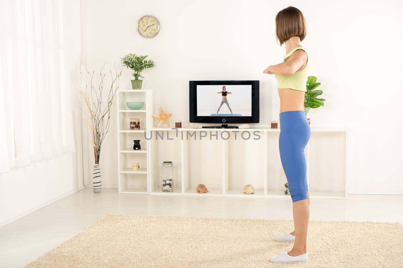Young woman in sports clothes, exercising in the room, watching the exercises on TV.
