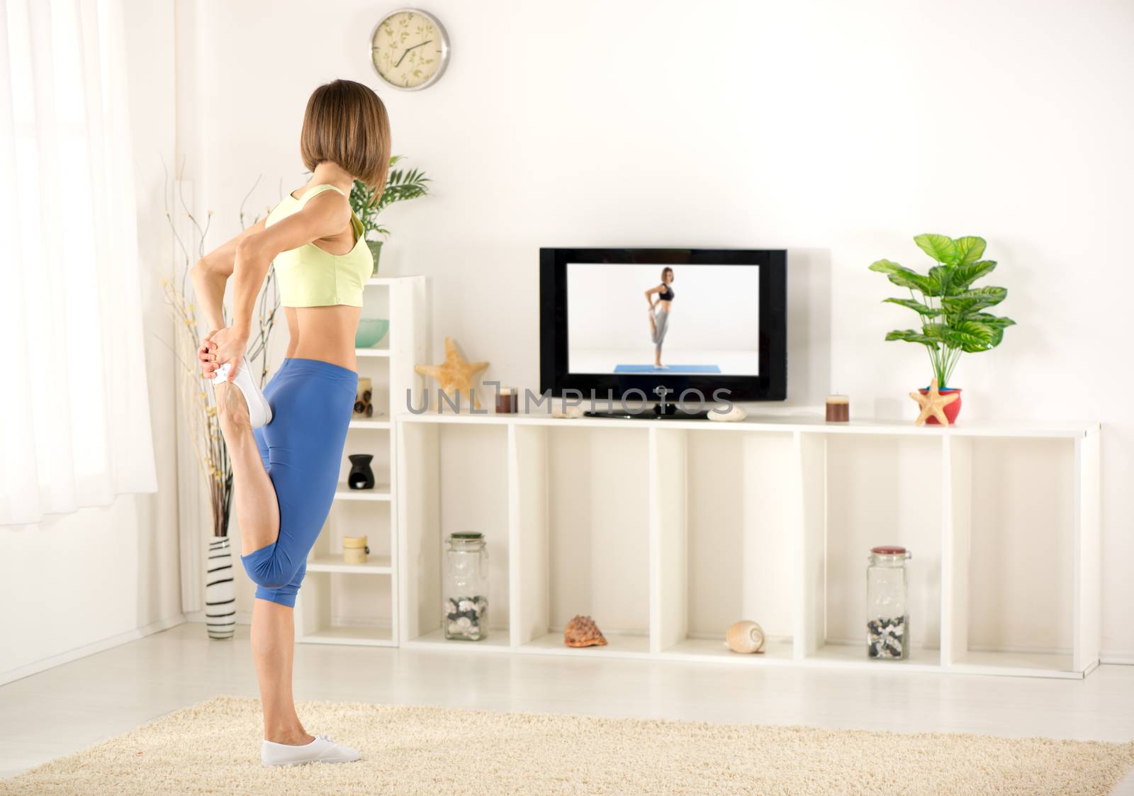Young woman in sports clothes, doing stretching exercises in the room, in front TV.