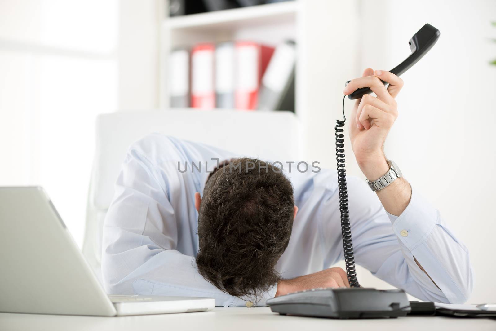 A young tired businesswoman, exhausted from work sleeping in front of his laptop, leaning on office desk with a telephone handset on hand.