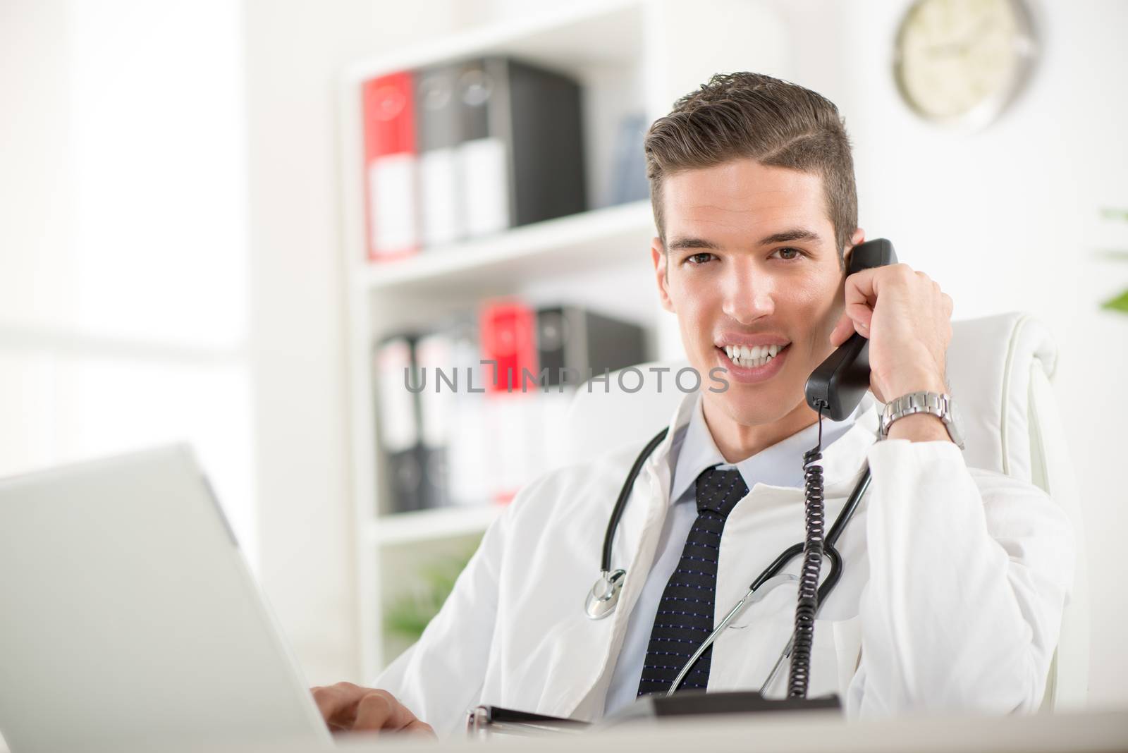 Young male doctor with stethoscope sitting at desk in doctor's room and phoning. Looking at camera.