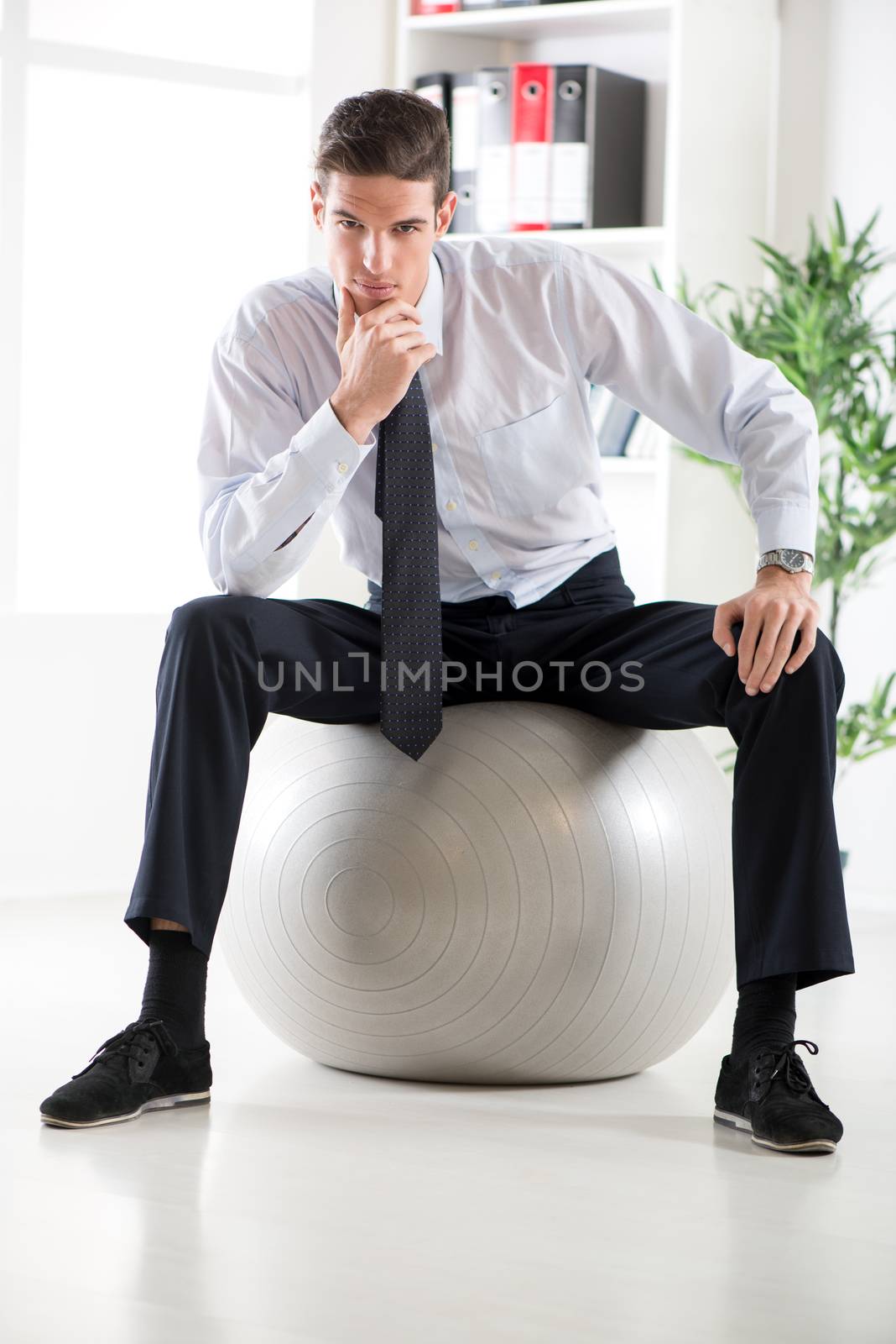 The young businessman sitting in the office on pilates ball and thinking. Looking at camera.