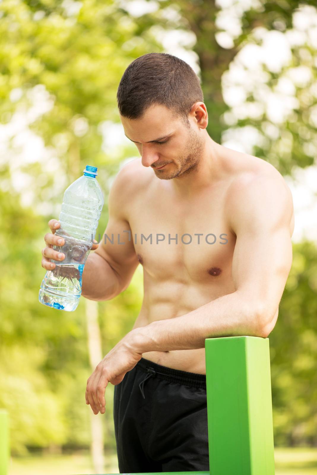 Naked muscle man resting after exercising in the outdoor gym. He is holding water bottle and thinking.