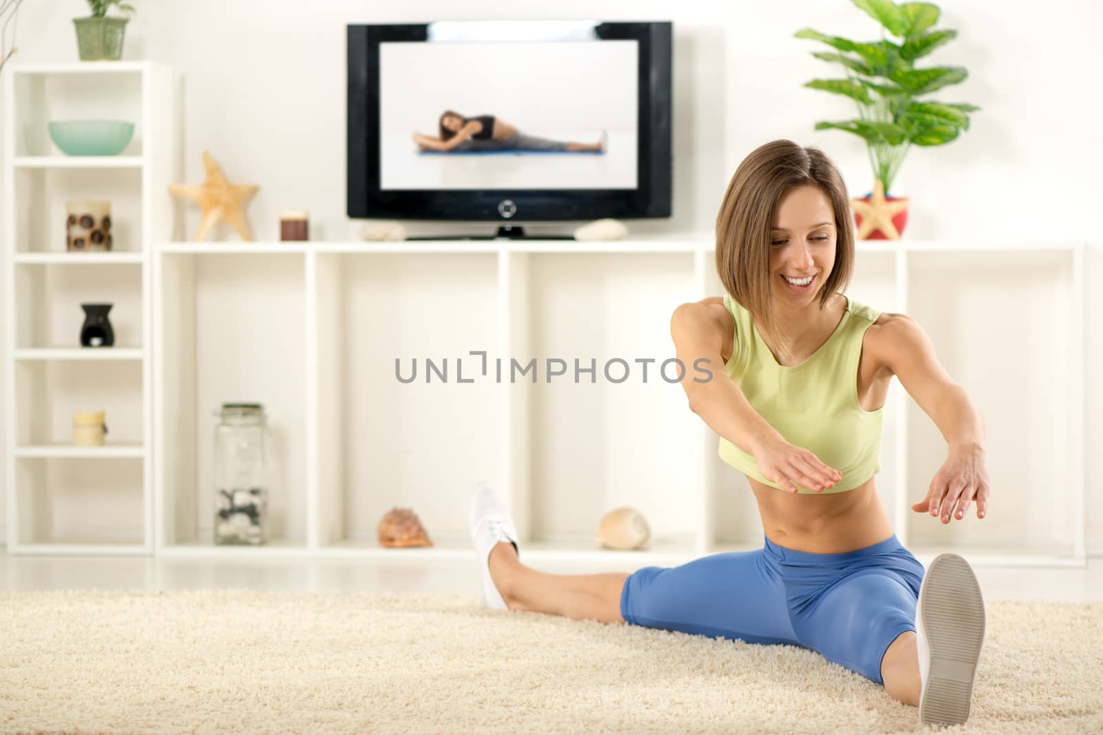 Young woman in sports clothes, doing stretching exercises in the room, in front TV.