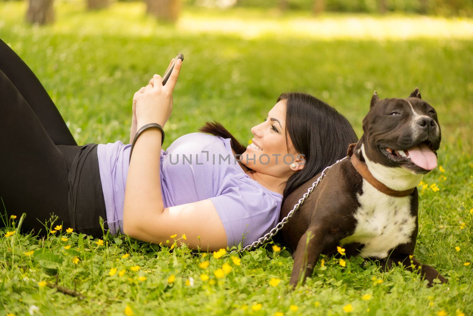Beautiful young woman lying on the her dog in the park and typing a message on your mobile.