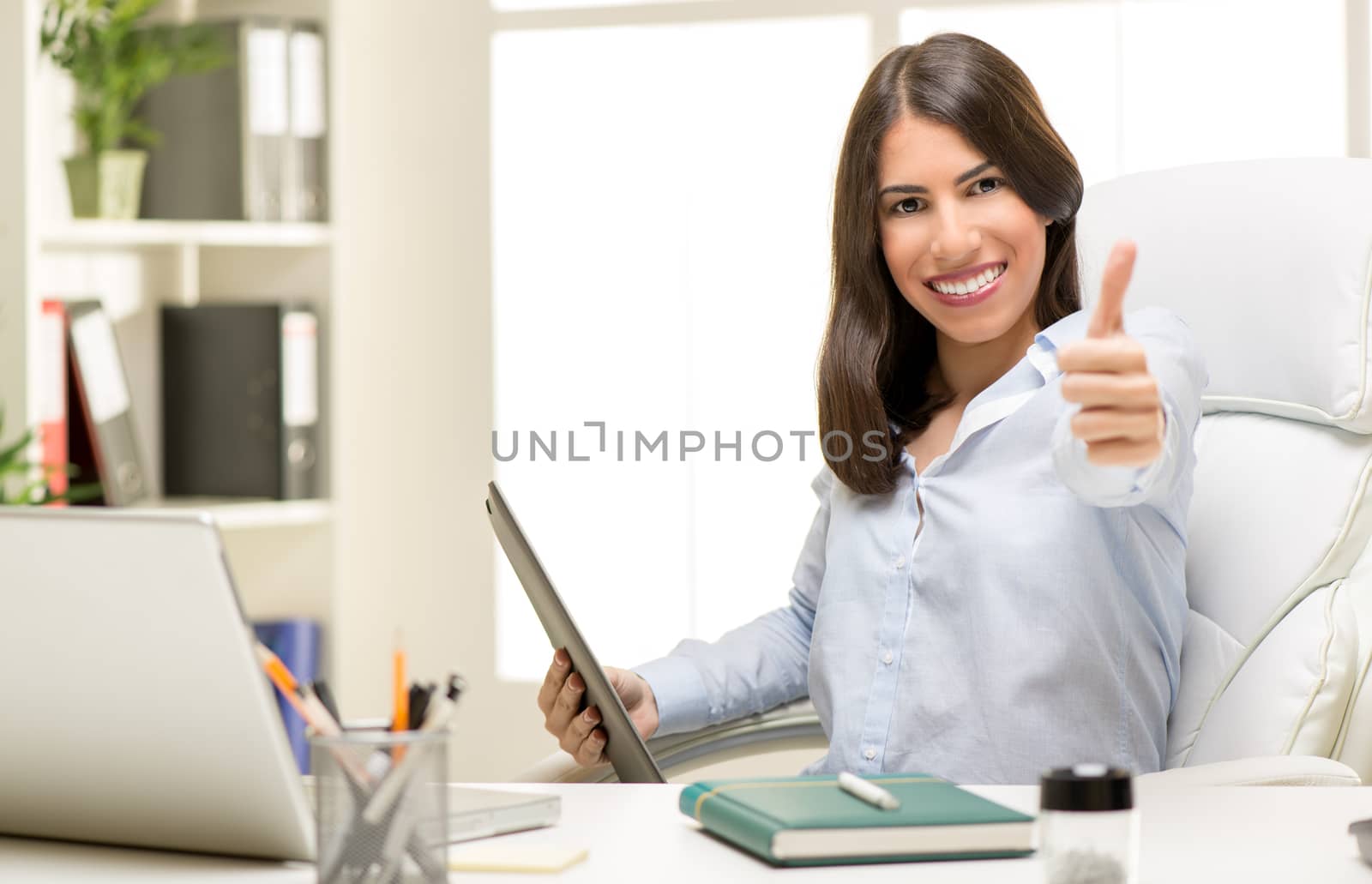 Successful businesswoman sitting at the desk in office with thumb up. Looking at camera and smiling.