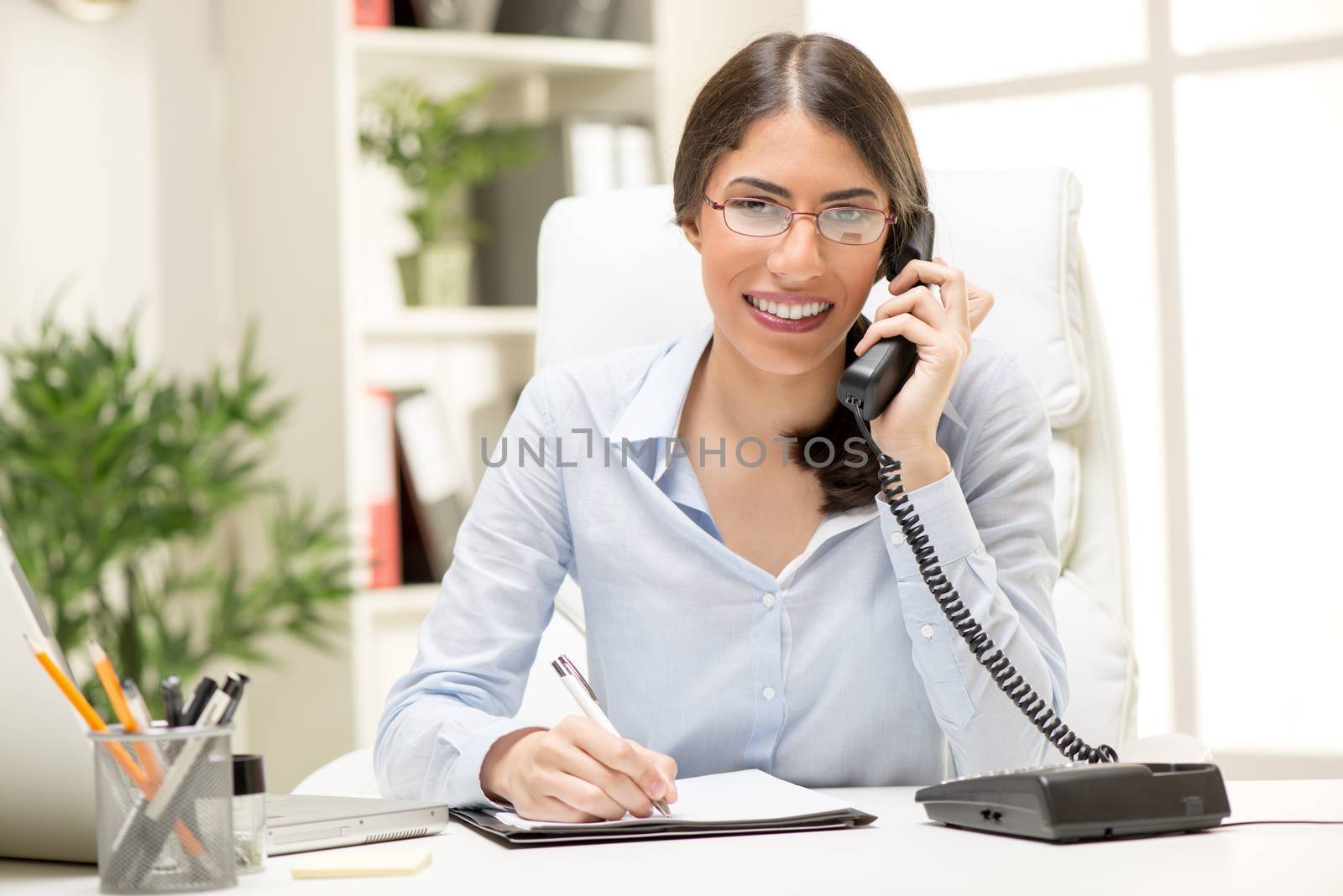 A young blond businesswoman phoning in office, sitting at an office desk in front of laptop, with a smile on her face looking at the camera.