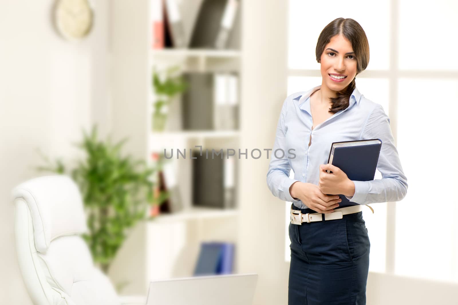 Beautiful young businesswoman standing in office, holding planner, smiling and looking at the camera. In the background you can see the shelves with binders.