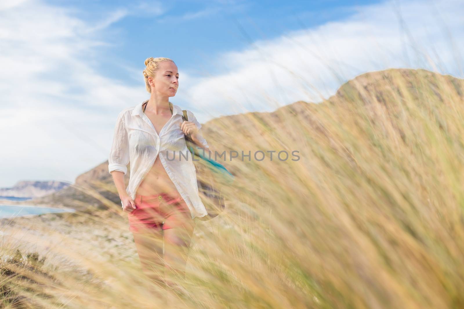 Relaxed woman on vacations in white loose shirt carrying beach bag and towel, enjoying beautiful coastline view of white sandy lagoon at Balos beach, Greece.