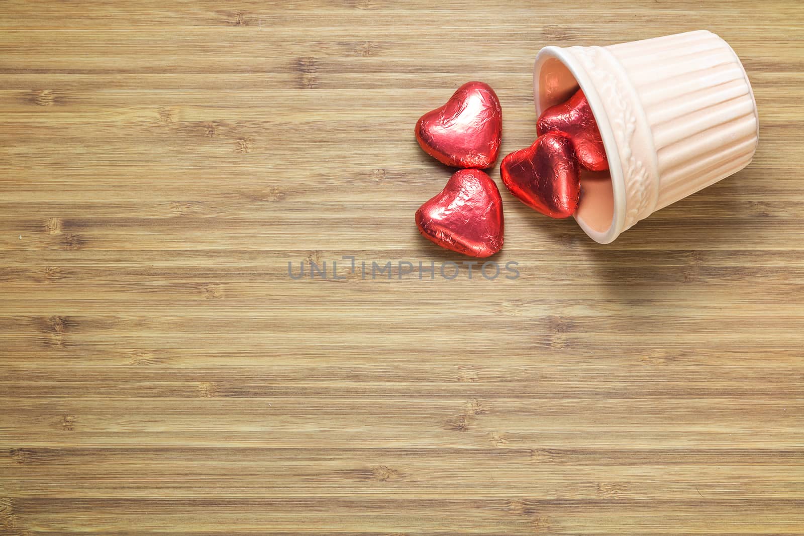 Heart shaped sweets wrapped in a bright red foil lying in a ceramic vase on a wooden texture. Background for romantic themes.