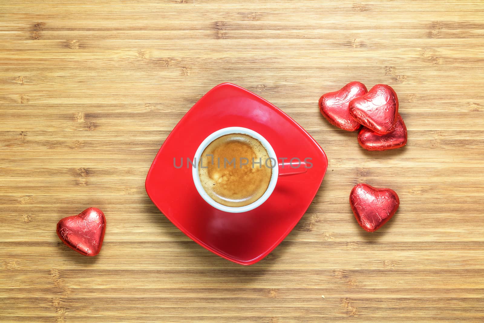 Heart shaped sweets wrapped in a bright red foil lying on a wooden texture with red cup of coffe near it. Background for romantic themes.