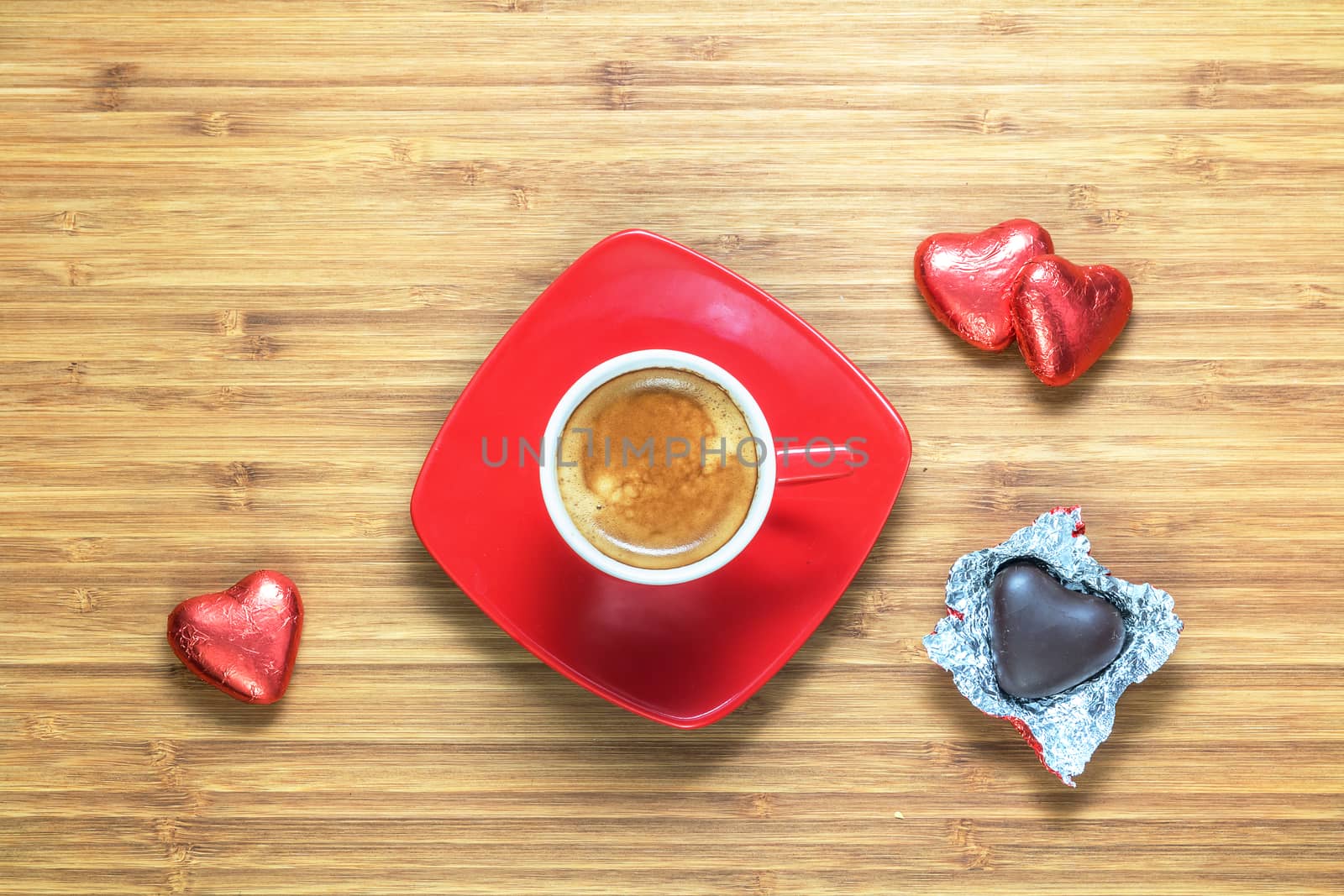 Heart shaped sweets wrapped in a bright red foil lying on a wooden texture with red cup of coffe near it. Background for romantic themes.