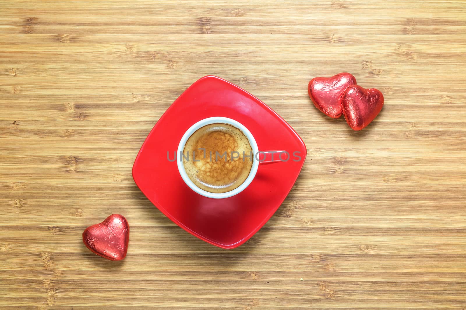 Heart shaped sweets wrapped in a bright red foil lying on a wooden texture with red cup of coffe near it. Background for romantic themes.