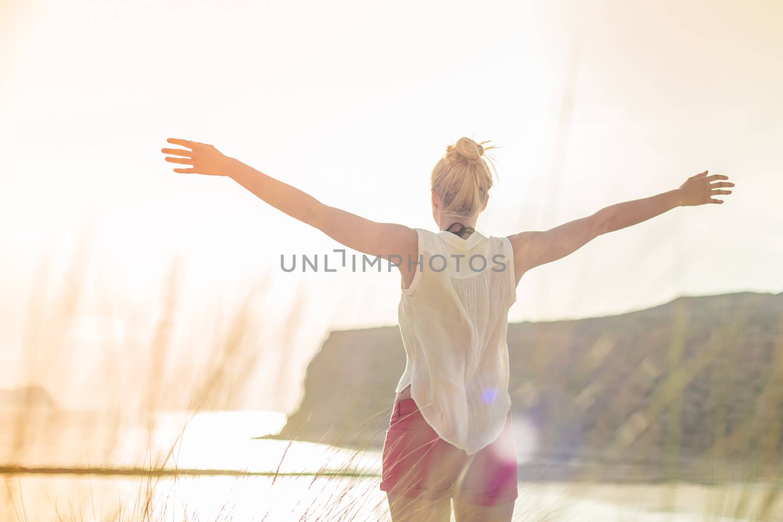 Relaxed woman in white shirt, arms rised, enjoying sun, freedom and life an beautiful beach. Young lady feeling free, relaxed and happy. Concept of vacations, freedom, happiness, joy and well being.