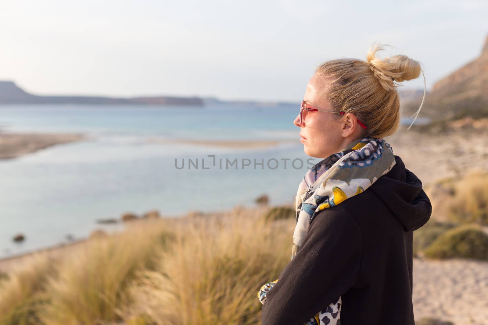 Relaxed woman in dark hoodie and colorful scarf looking at distance, enjoying beautiful nature, freedom and life at serene landscape at Balos beach, Greece. Concept of well being.