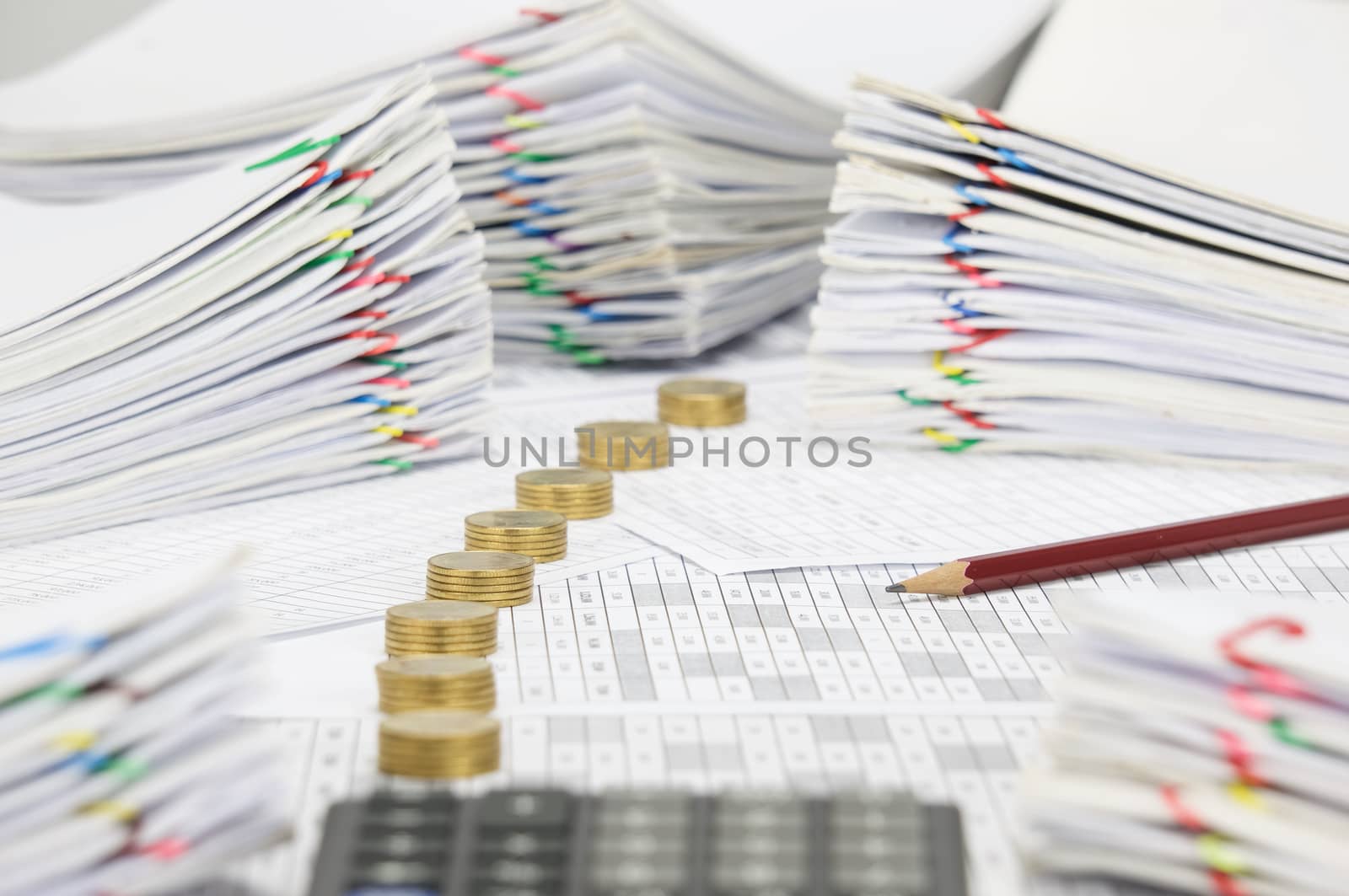 Brown pencil on finance account have blur calculator and pile line of gold coins and pile overload paperwork of report and receipt with colorful paperclip place as foreground and background.