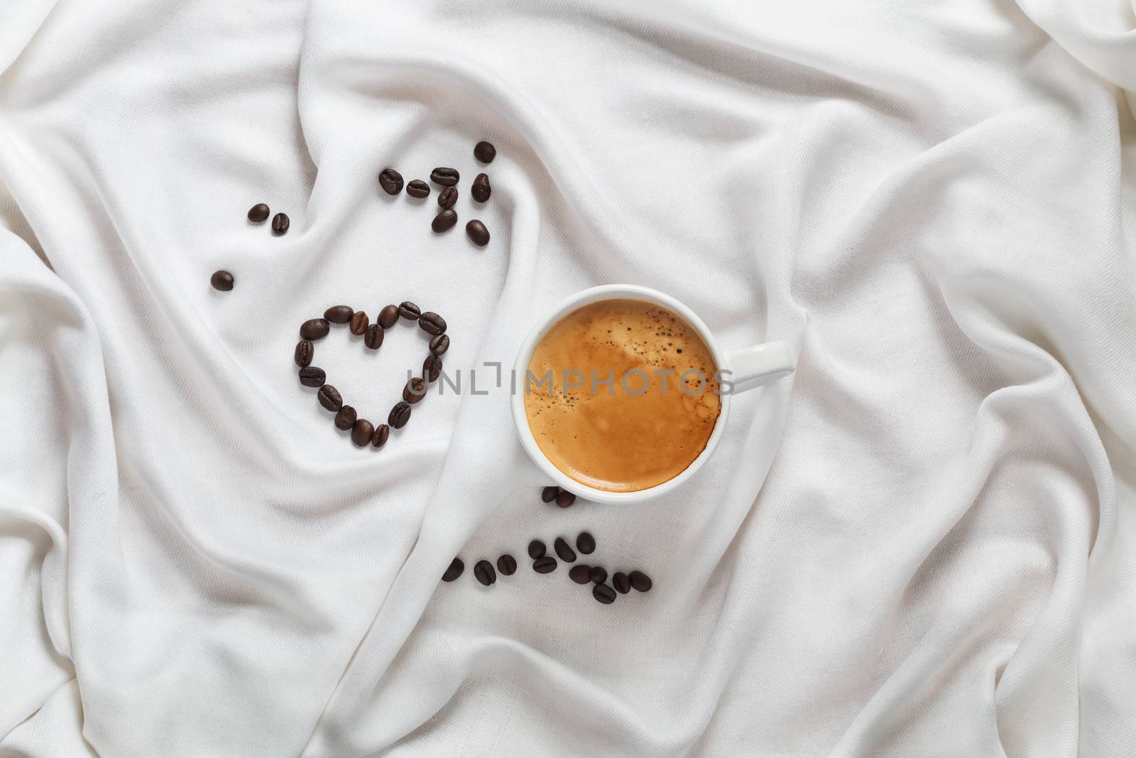 Cup of coffee on a white silk fabric. Espresso and scattered beans in the form of a heart. Top view. Romantic breakfast for Saint Valentine's Day.