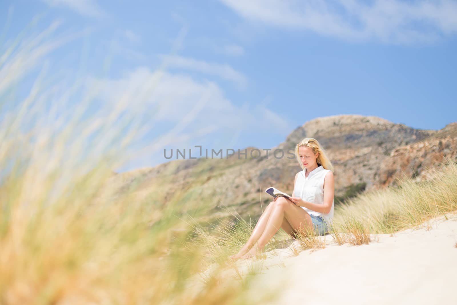 Relaxed woman enjoying sun, freedom and good book an beautiful sandy beach of Balos in Greece. Young lady reading, feeling free and relaxed. Vacations, freedom, happiness, enjoyment and well being.