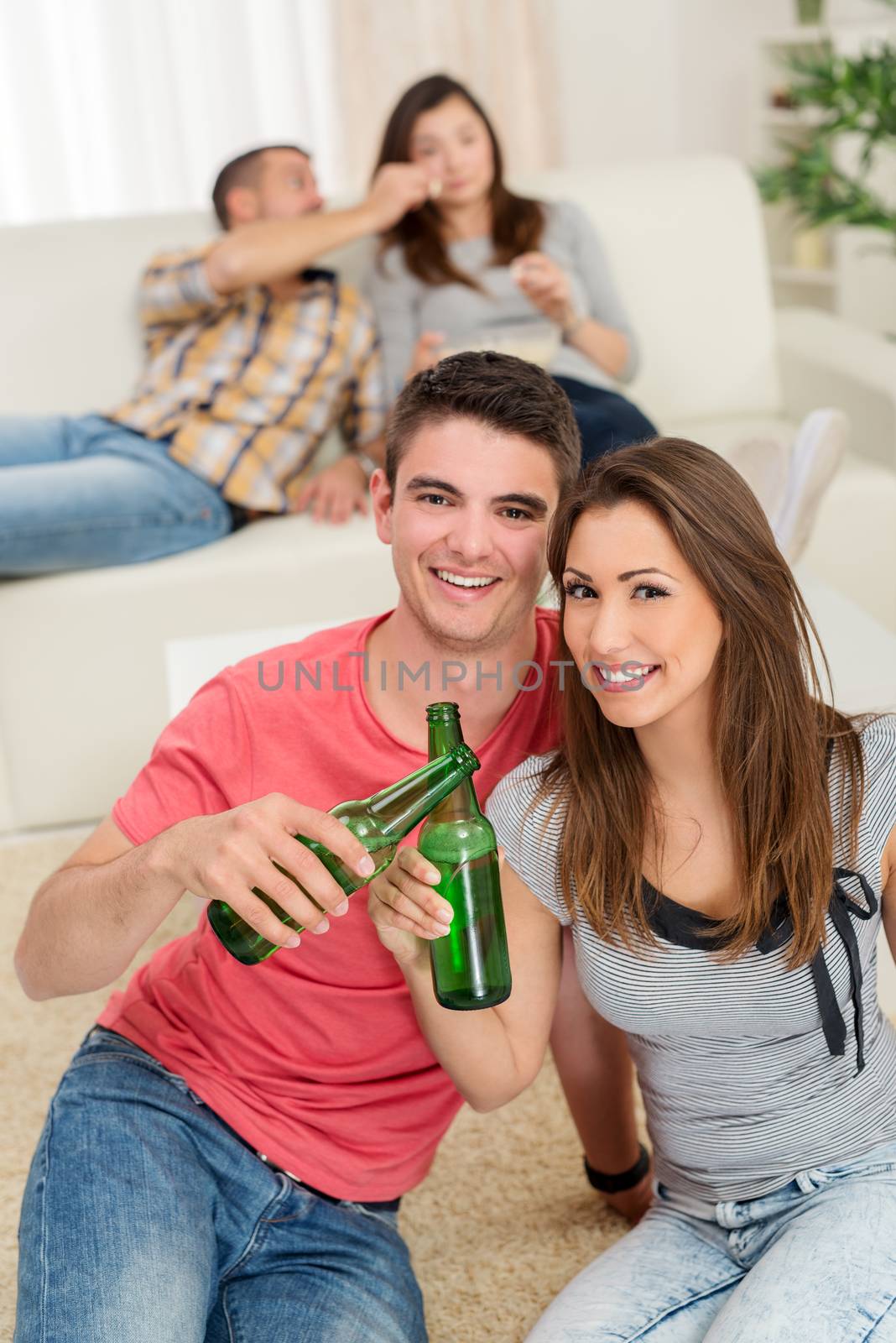 Young happiness couple enjoying at home party and toasting with beer. Their friends in the background. Selectiv focus. Focus on foreground. Looking at camera.