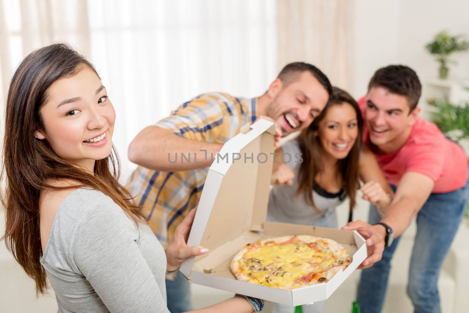 Four cheerful friends enjoying pizza together at home party. Selective focus. Focus on foreground, on Japanese girl. She is holding box with pizza and looking at camera.