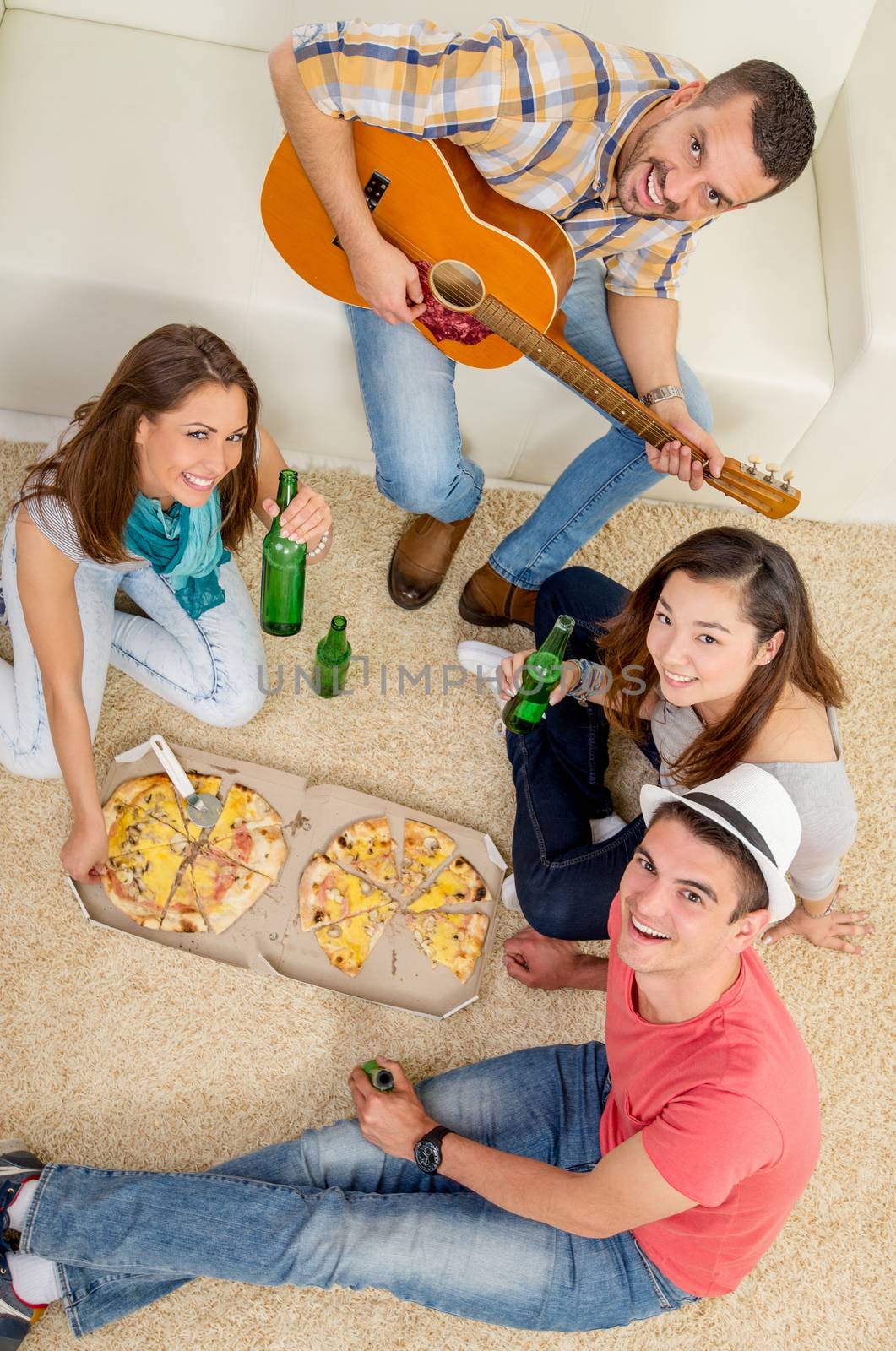 Four cheerful friends enjoing with guitar in an apartment. They drinking beer and eating pizza. Looking at camera.
