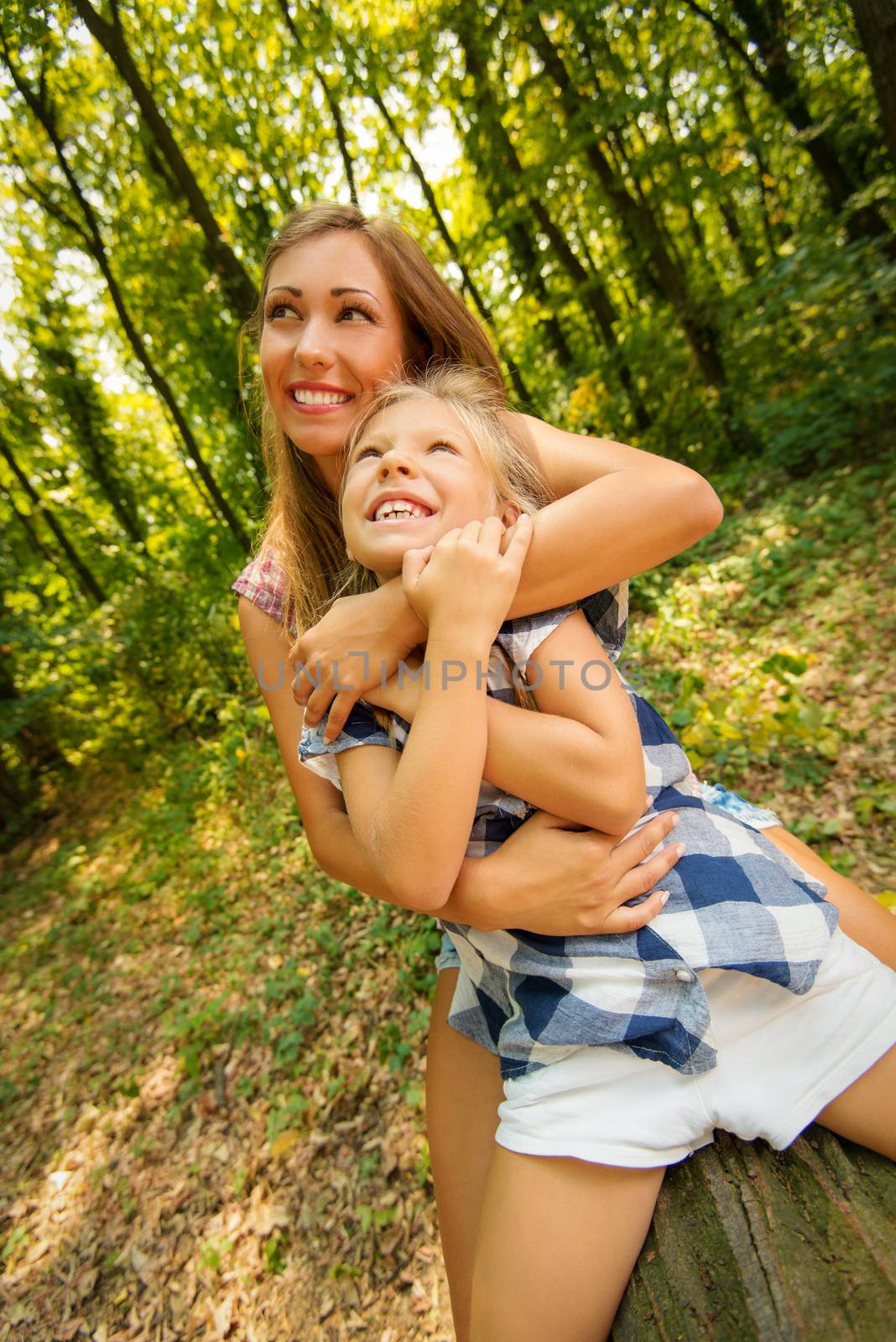 Beautiful young mother and her daughter sitting on fallen tree in the forest and enjoying. 