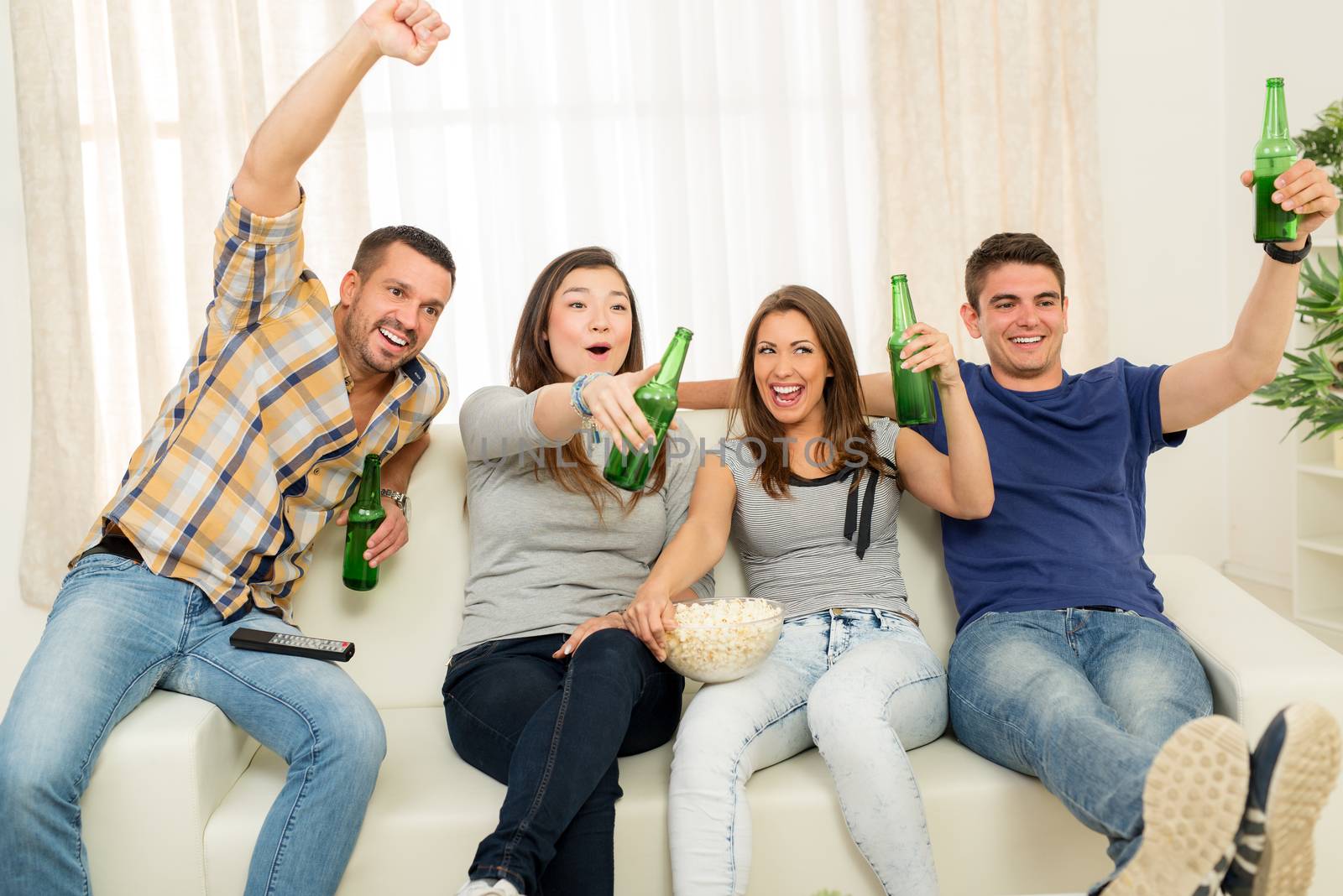 Four cheerful friends hanging out in an apartment and watching a football game together. They cheering, drinking beer and eating popcorn.